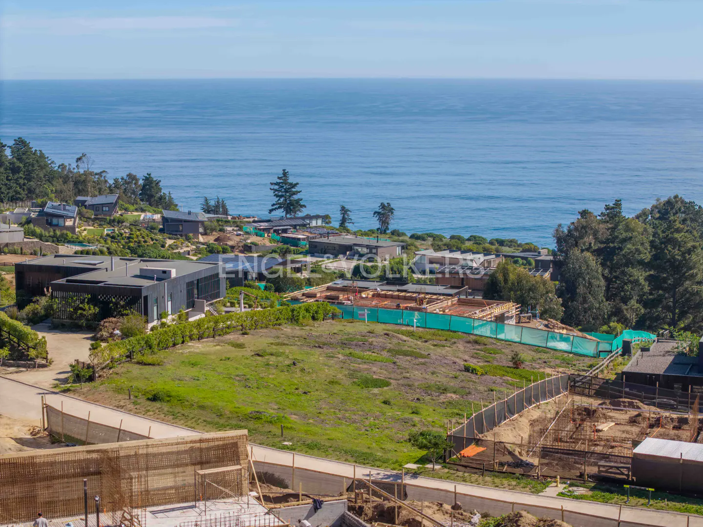 Ocean view property with modern homes and construction. Green grass, trees, and blue ocean under a clear sky. Engel & Volkers logo visible.