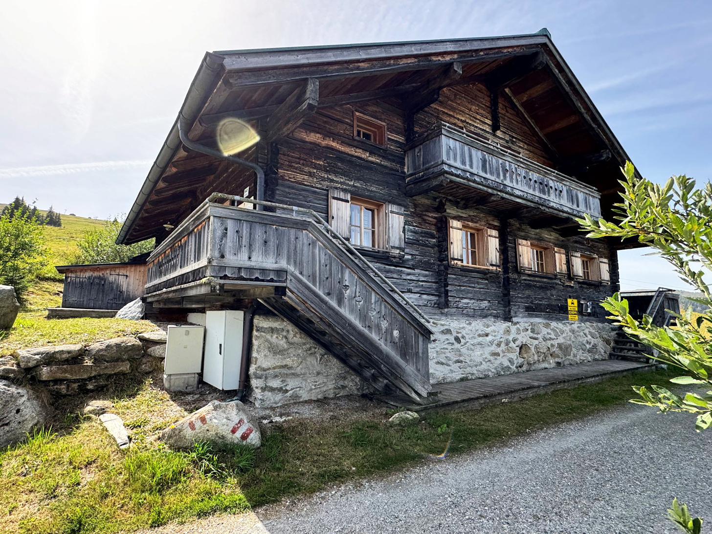 A rustic, dark wood Alpine cabin with a stone foundation, wooden balcony, and stairs, set against a grassy mountain hillside. [AI-generated text]