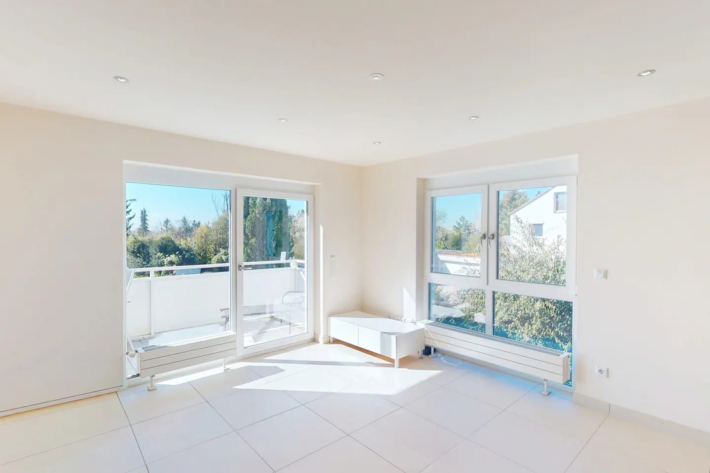 Bright, empty room with white tile floor, white walls, and large windows. Balcony visible through sliding glass doors.