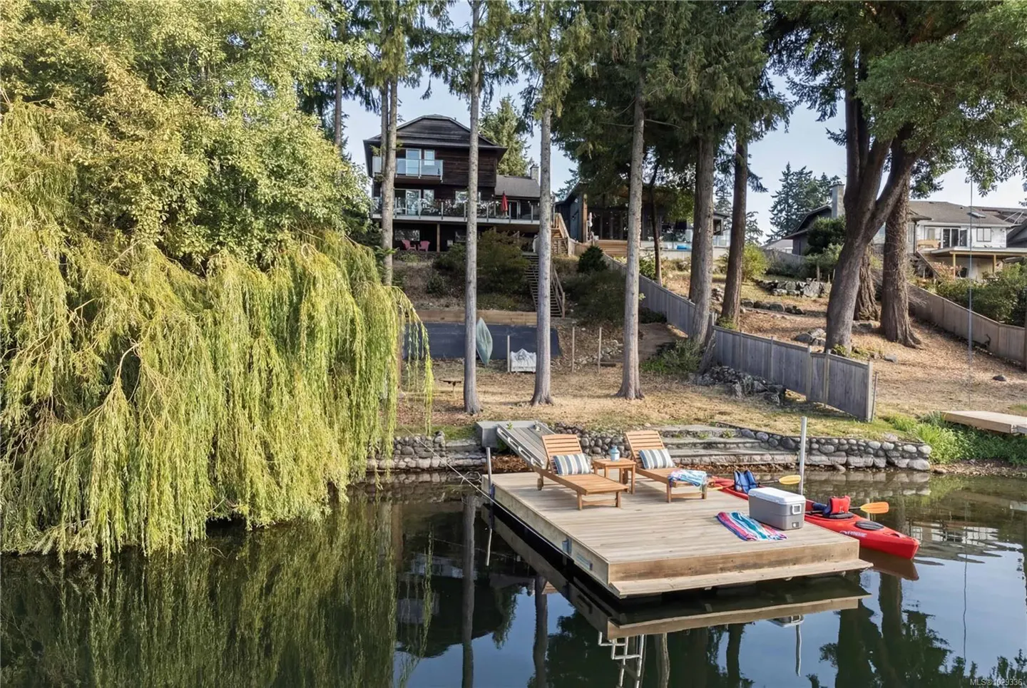 Waterfront property with a wooden dock, lounge chairs, kayaks, and a two-story house on a hill surrounded by trees.