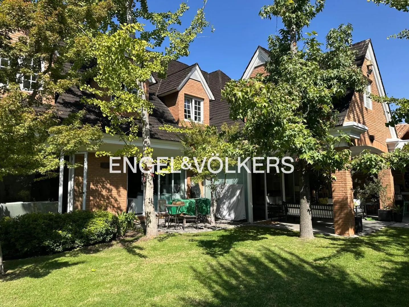 Exterior view of a two-story brick house with a dark roof and green lawn, partially obscured by trees. Engel & Volkers logo overlay.
