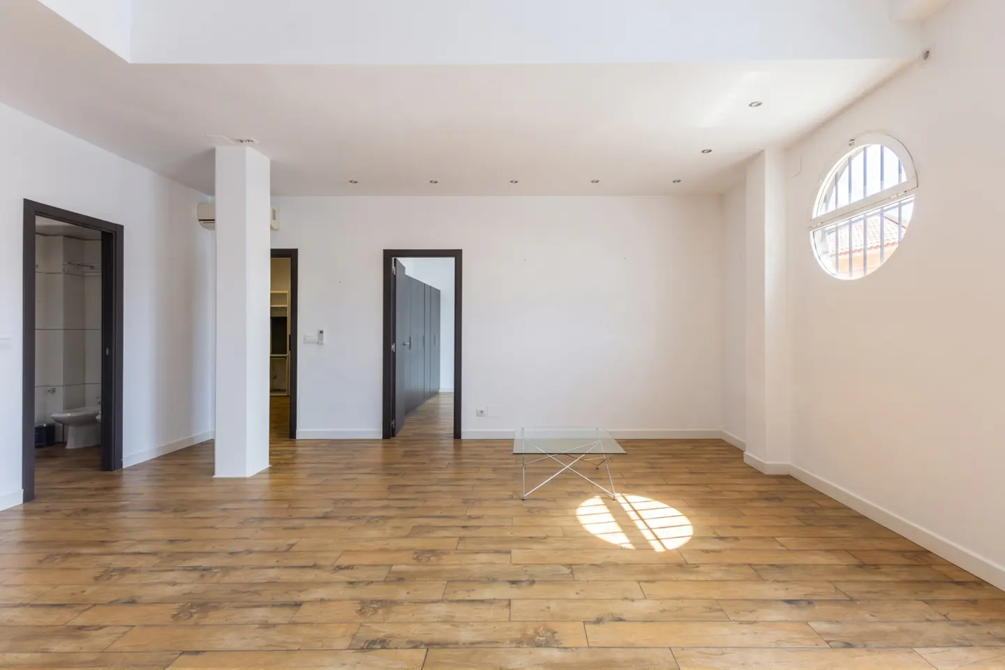Bright, empty room with wood floors, white walls, and two dark-framed doorways. A round window with bars casts sunlight on the floor.