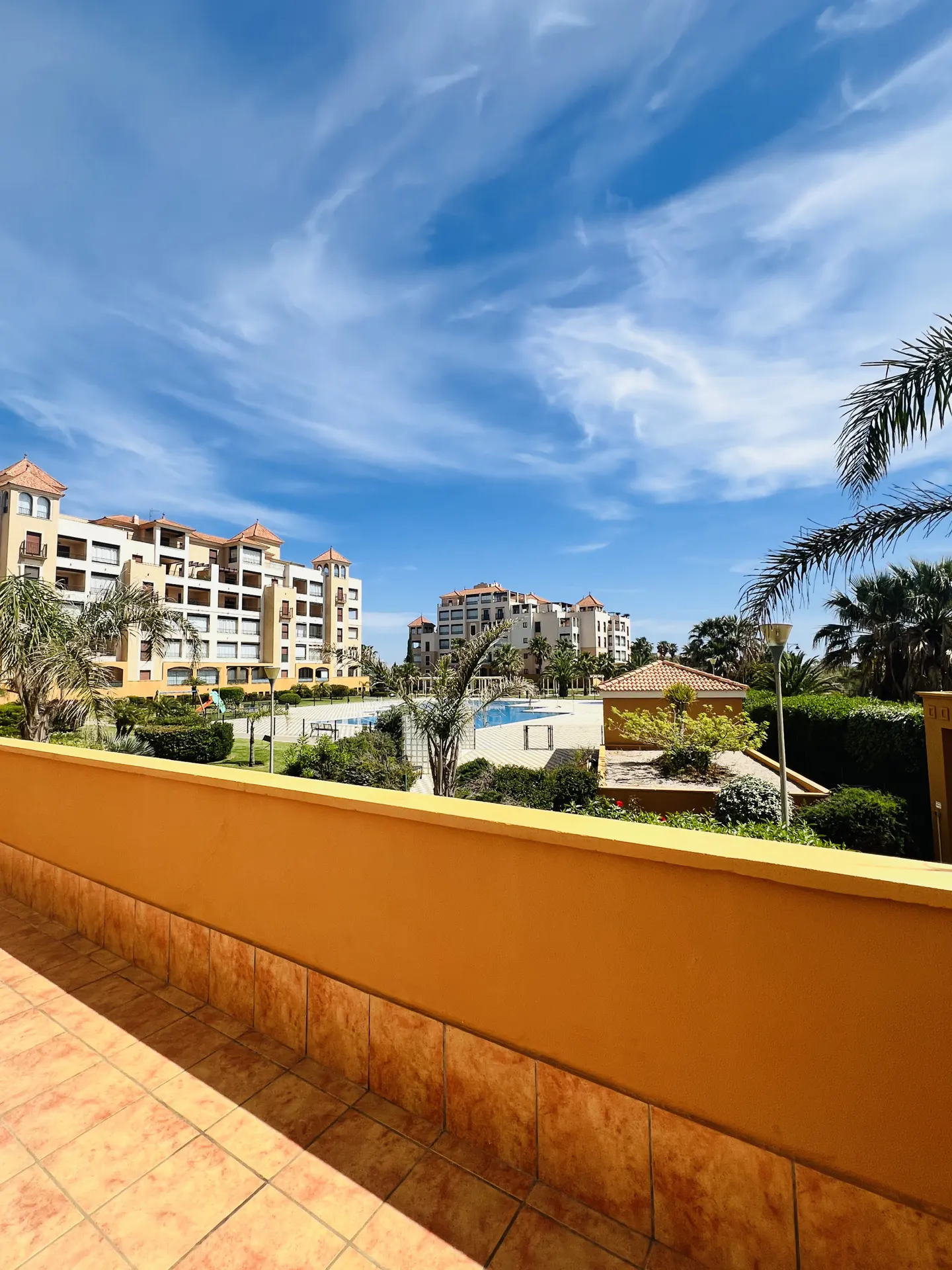 View from a balcony with orange walls overlooking a pool and apartment buildings under a blue, cloudy sky.
