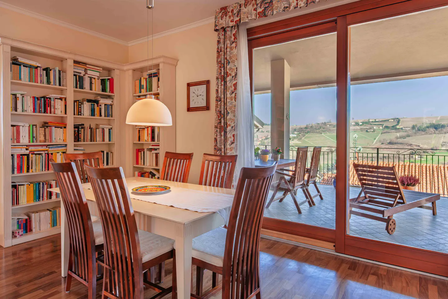 Dining room with a white table, wooden chairs, and a bookshelf. Sliding doors open to a balcony with a view of rolling hills.