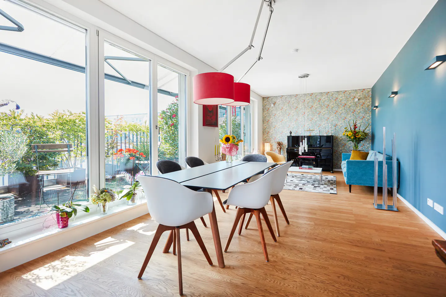 Bright, modern dining room with a white table, chairs, and red pendant lights. Large windows overlook a green balcony. A blue sofa and piano are in the background.