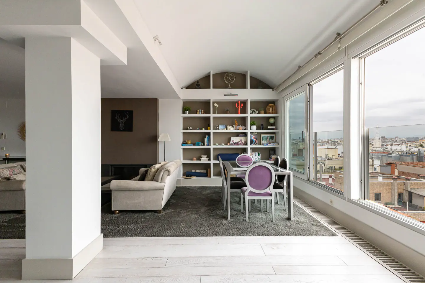 Bright, modern living room with a gray rug, white walls, and a large window overlooking a city. A dining table with purple chairs sits near a built-in bookshelf.