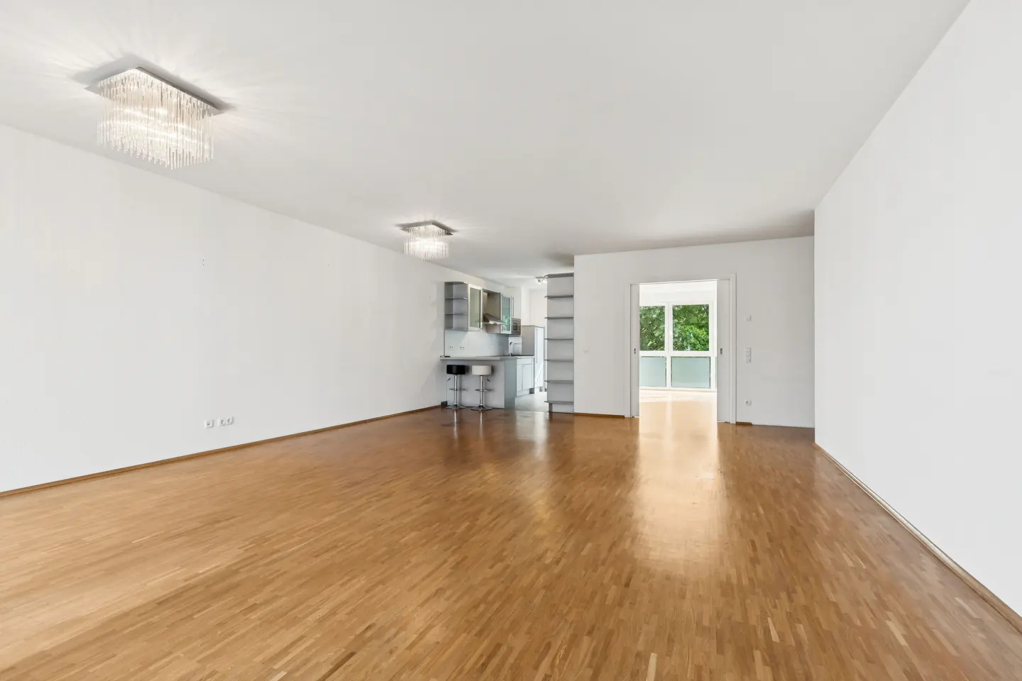 Spacious, bright living room with light wood floors, white walls, modern crystal ceiling lights, and an open kitchen area.