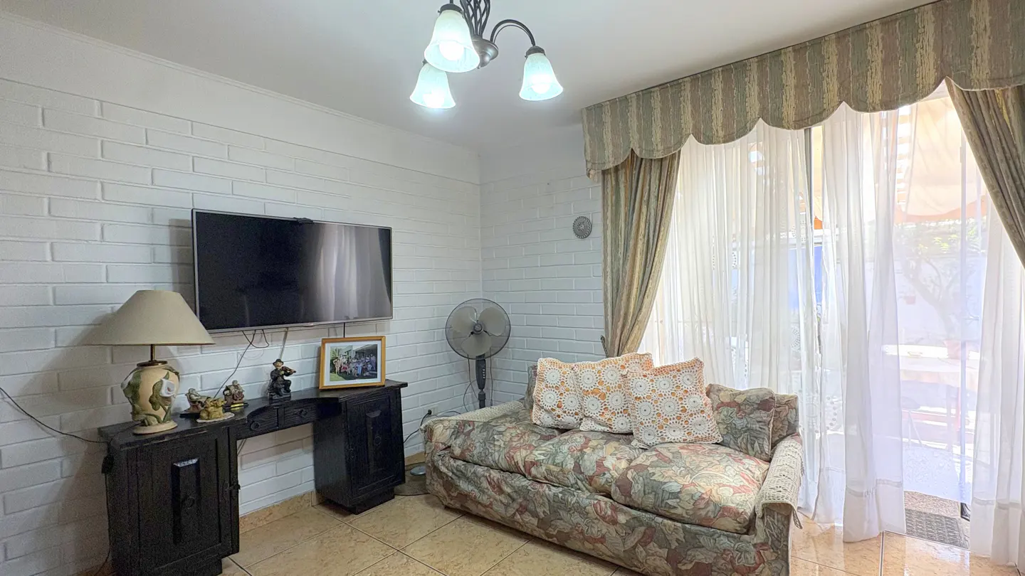 Living room with white brick walls, floral sofa, TV, and dark wood console table. Sheer curtains cover a bright window.