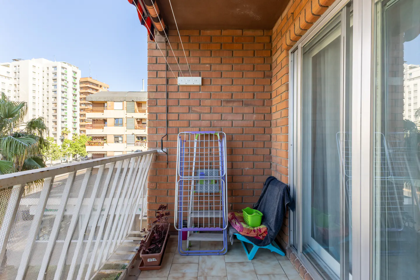 A balcony with a brick wall, white railing, and a view of buildings. A clothes drying rack and a blue chair are also visible.