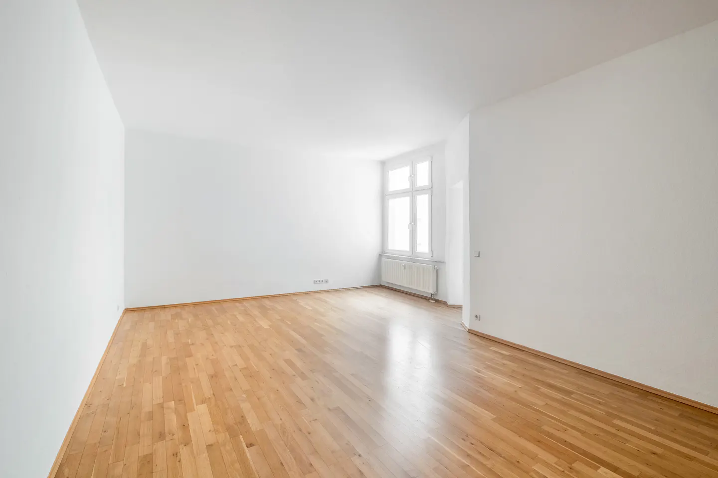 Bright, empty room with hardwood floors, white walls, and a large window with a radiator below.