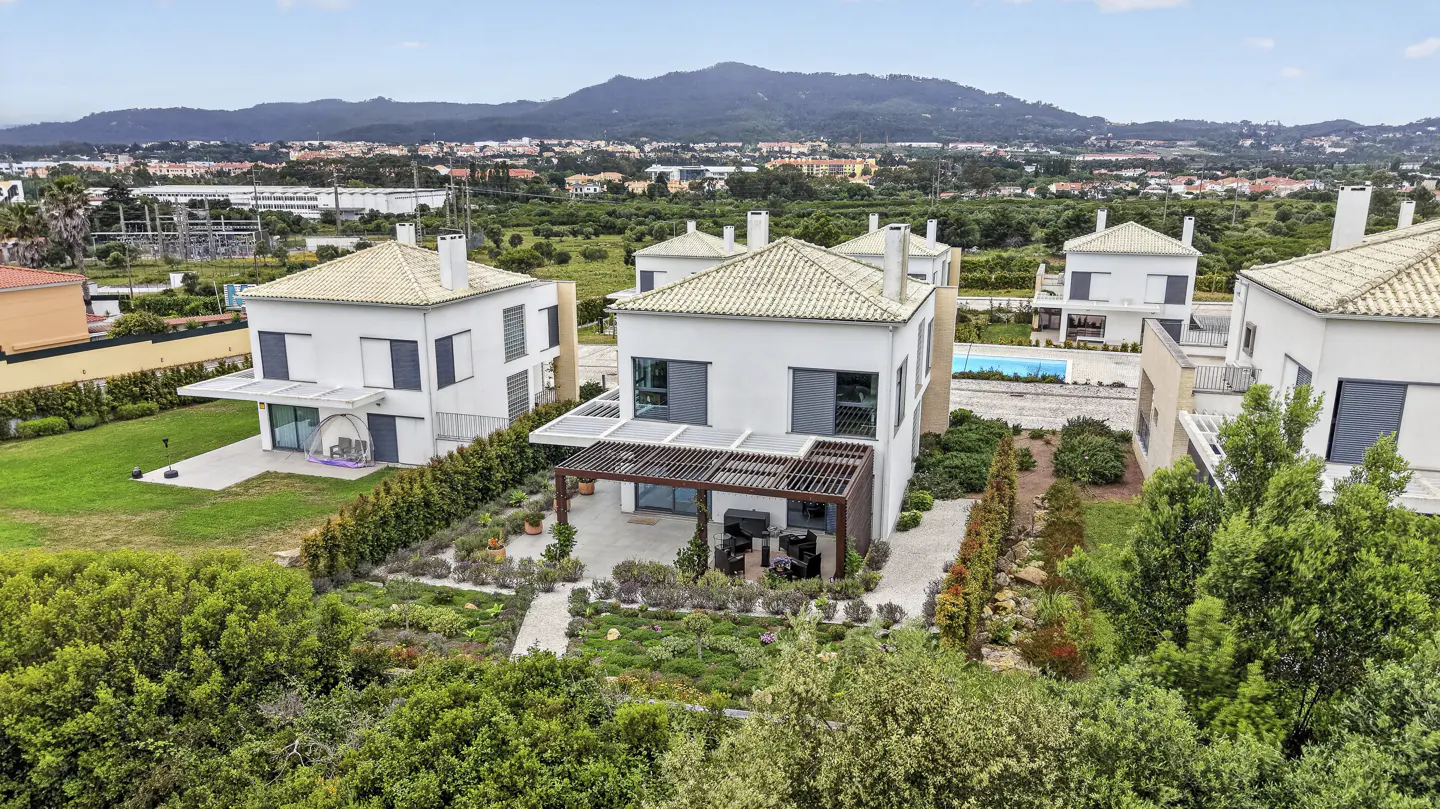 Aerial view of modern white houses with beige tile roofs, green lawns, and a mountain backdrop.