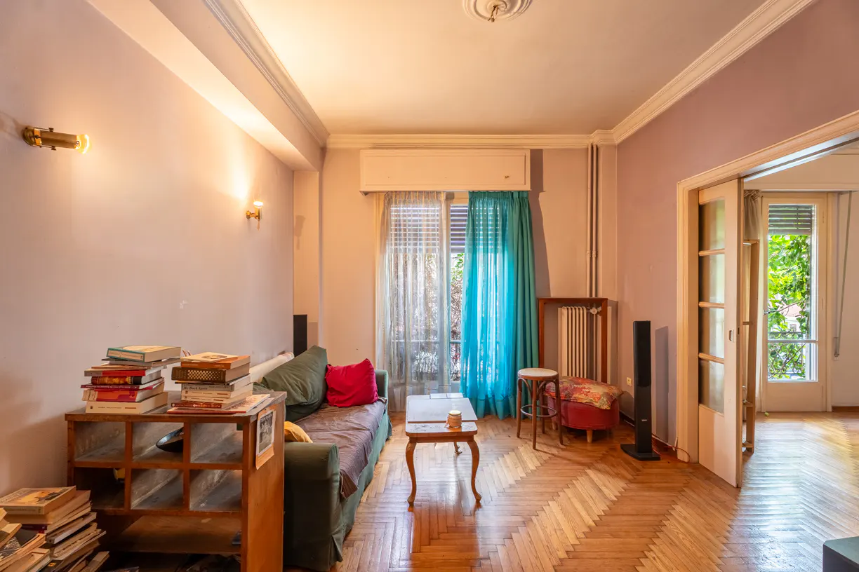 Living room with parquet floors, a green sofa with a red pillow, and a wooden bookshelf stacked with books. Turquoise curtains hang by the window.