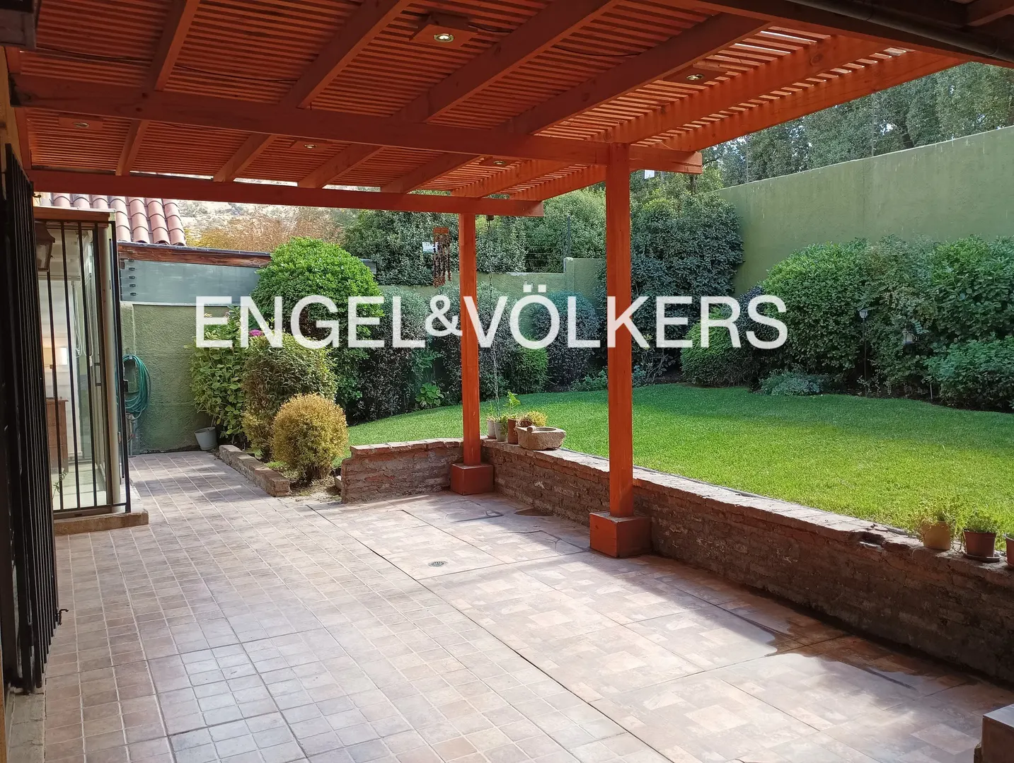 Outdoor patio with orange pergola, brick wall, and tiled floor. Green lawn and bushes in the background. Engel & Völkers logo.