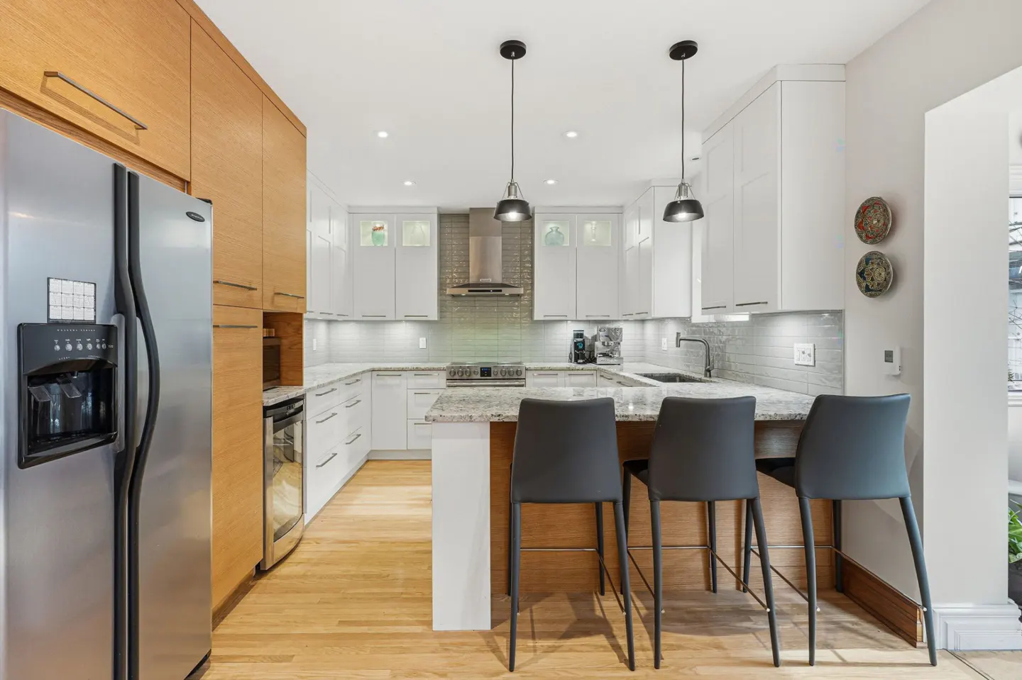 A modern kitchen with white cabinets, granite countertops, and stainless steel appliances. Three black bar stools face the island.