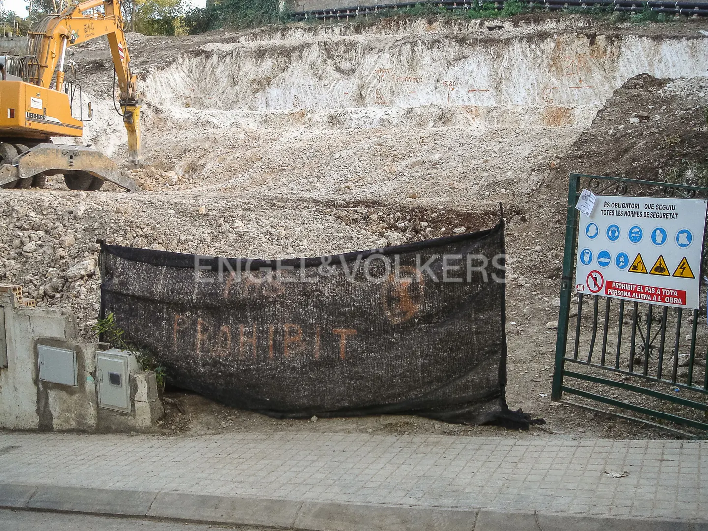 Construction site with a yellow excavator breaking ground. A black mesh banner with "Engel & Volkers" and "Prohibit" is in the foreground. A safety sign is on a green gate.