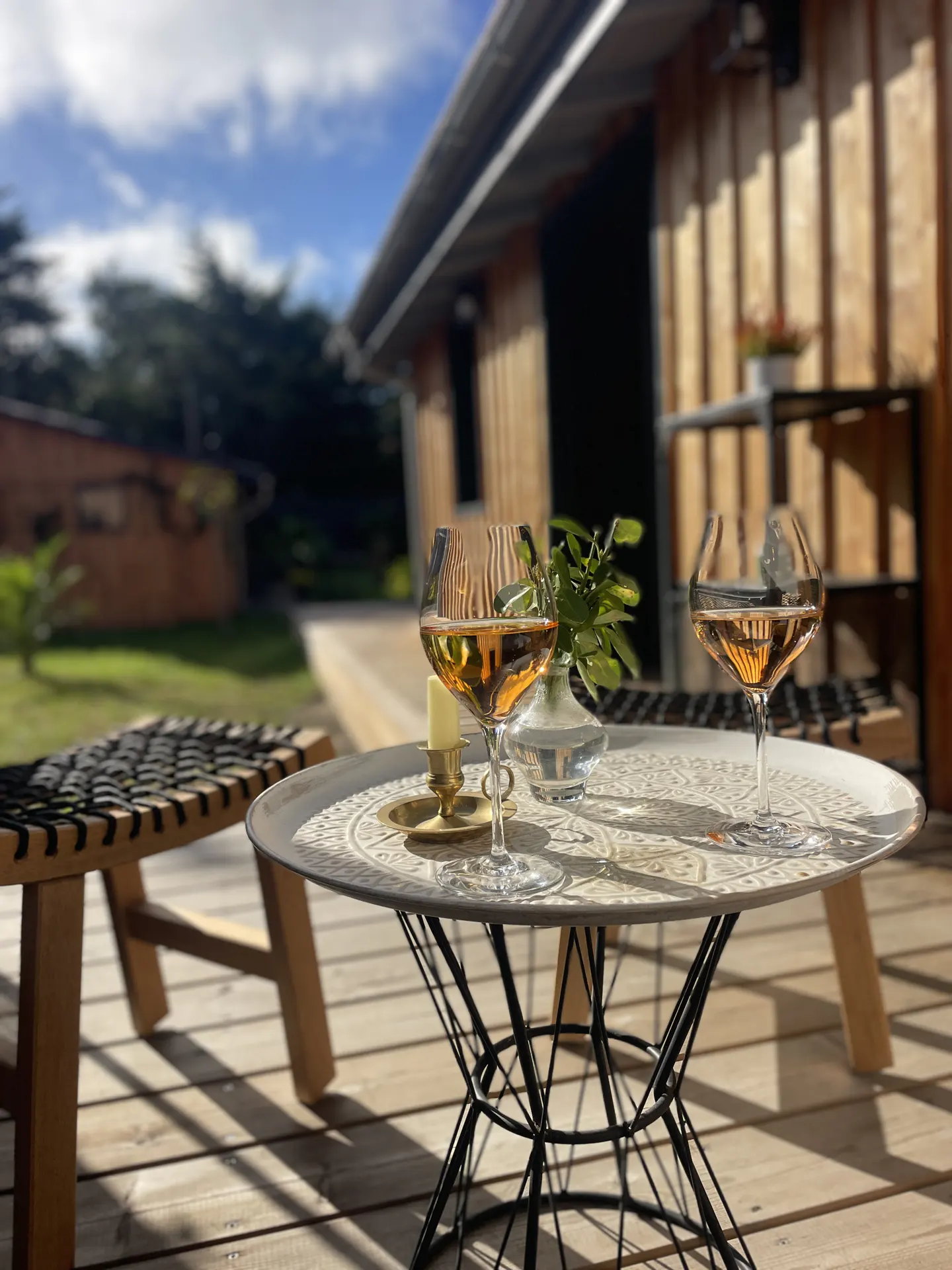 Two wine glasses on a table outside a wooden cabin. A bench is nearby, and trees are visible in the background.