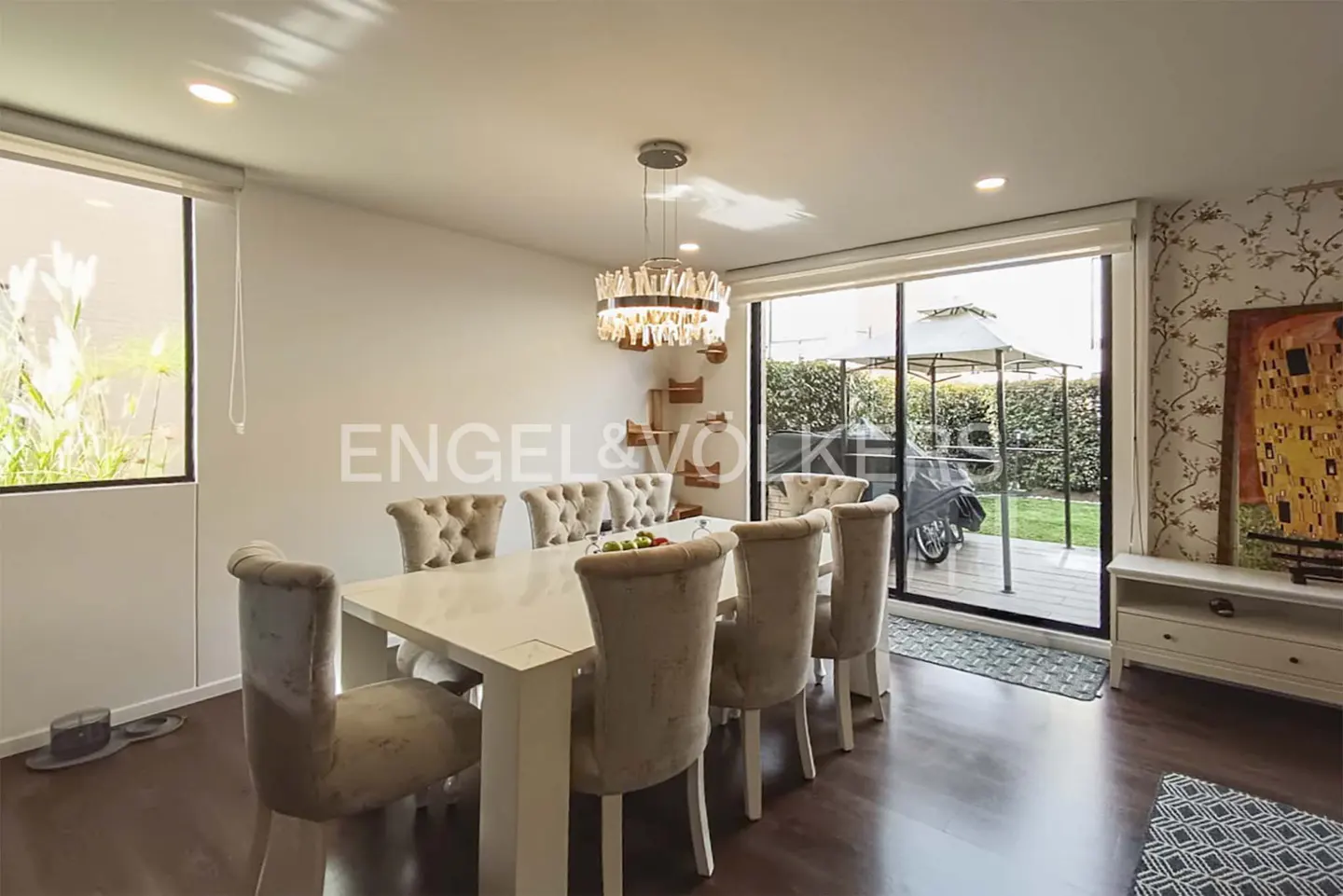 Bright dining room with a white table, beige chairs, and a crystal chandelier. A sliding glass door leads to a patio and garden.