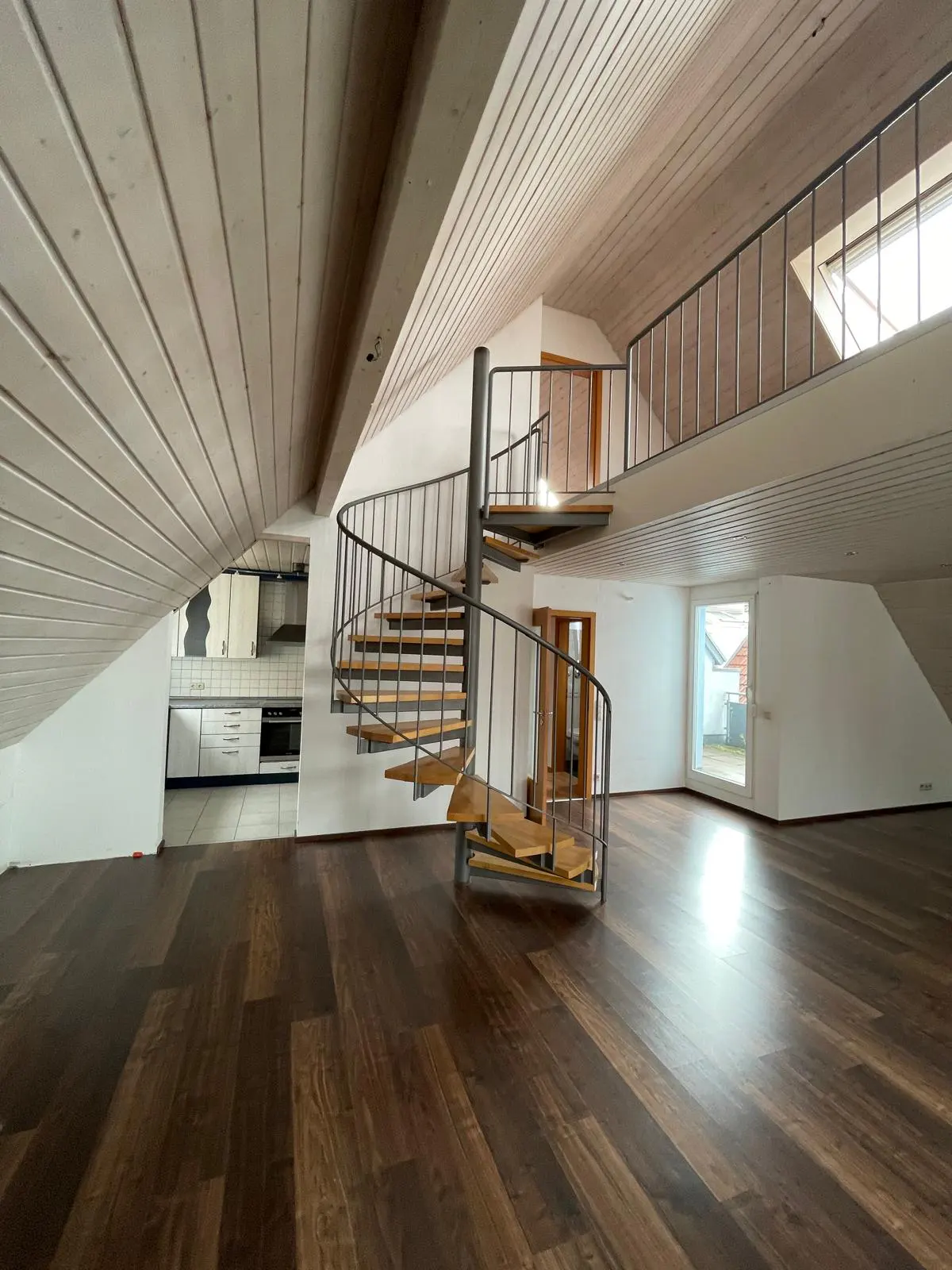 Spacious attic room with dark wood floors, a metal spiral staircase, and a view into a modern kitchen.