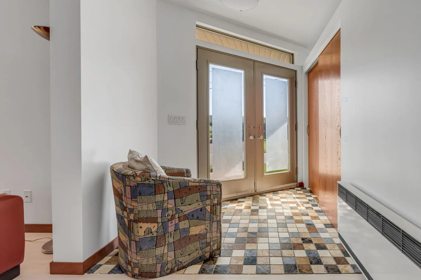 A patterned armchair sits on a multi-colored tiled floor in a bright entryway with double glass doors and wood closets.