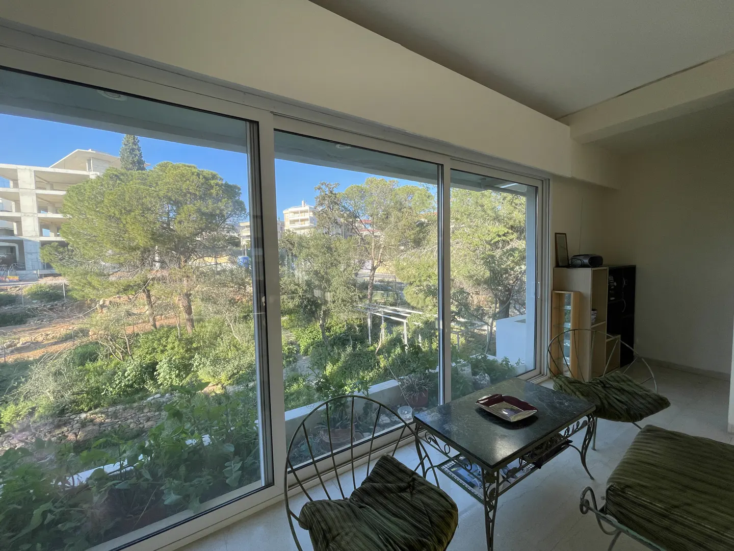 Living room with a view of lush greenery through large windows. Metal chairs and table with a dark top. Neutral wall color.