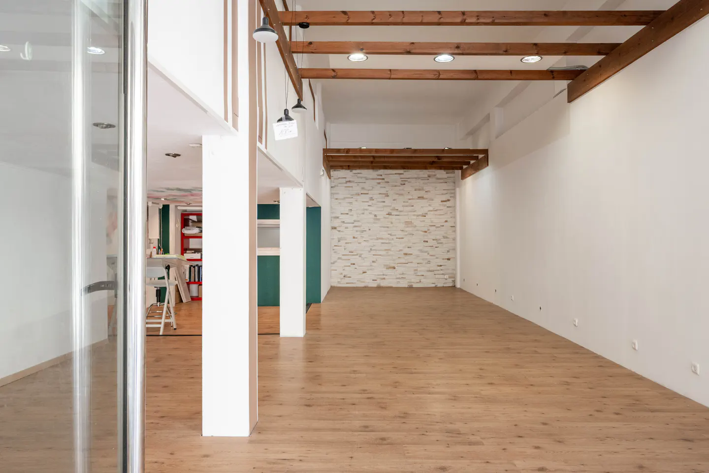 Bright, empty room with wood floors, white walls, and exposed wood beams on the ceiling. A brick accent wall is visible in the back.