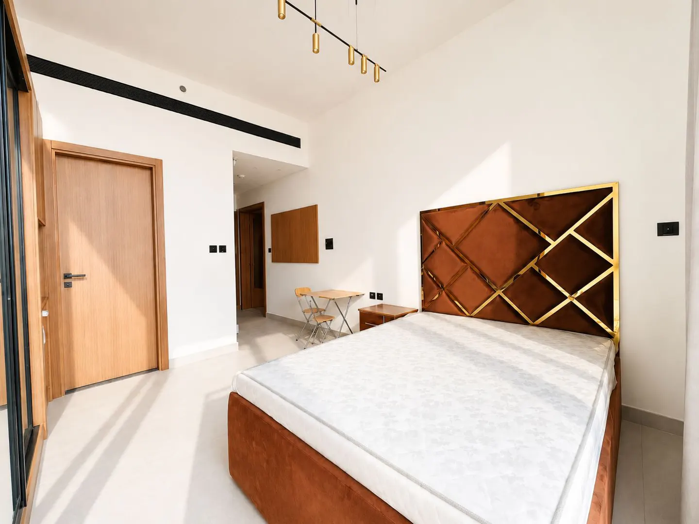Bedroom with a brown bed and gold headboard, white walls, and a small table with chairs in the background.