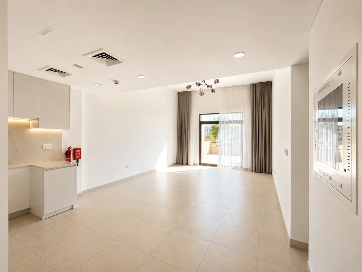 Bright, open-plan living area with white walls, tiled floors, a modern kitchenette, and a balcony with floor-length curtains.