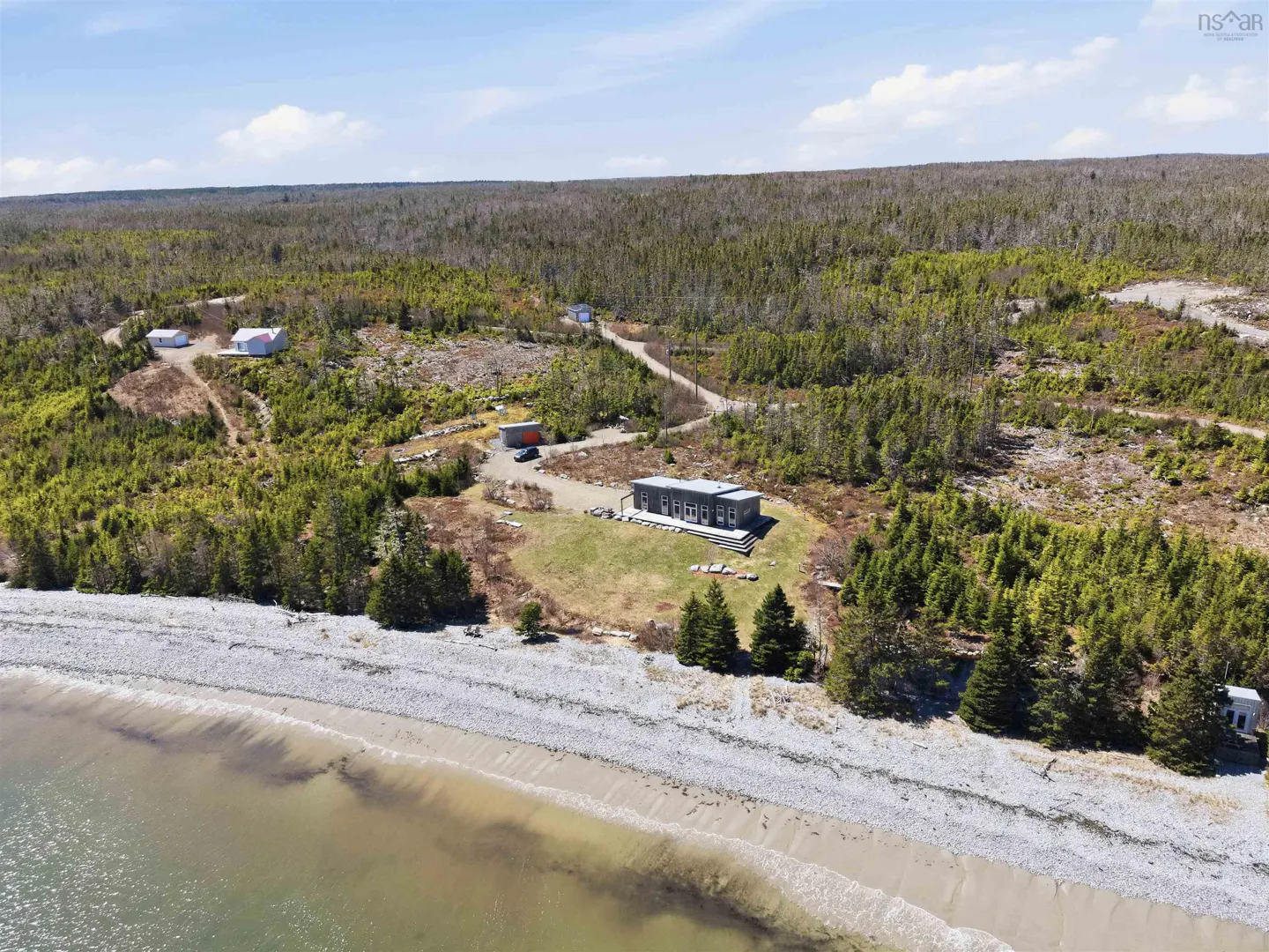 Aerial view of a modern grey home on a grassy lot by a rocky beach, surrounded by dense forest and smaller outbuildings.