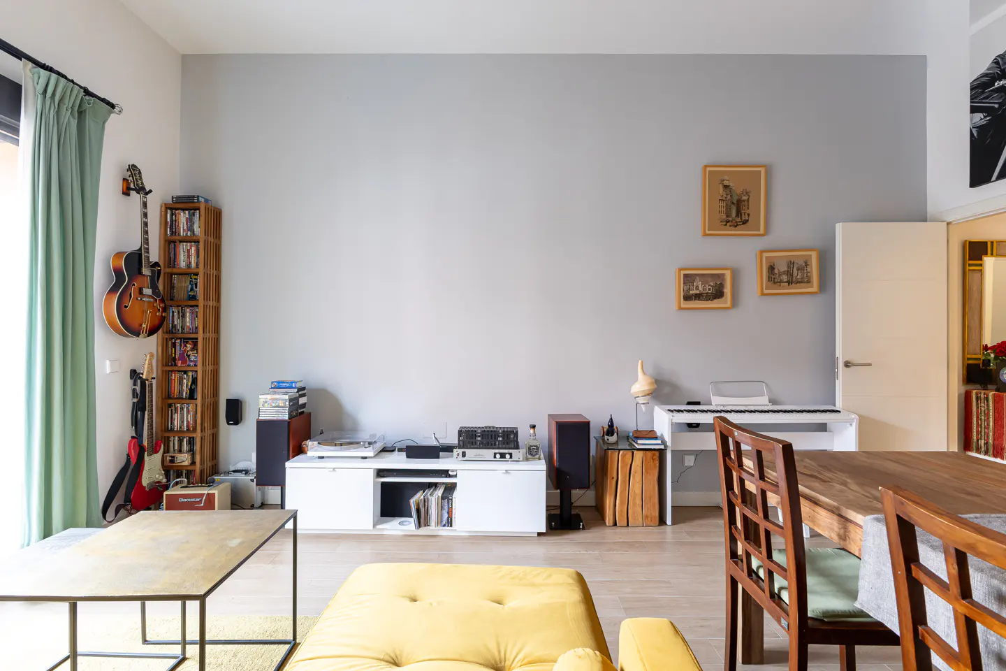 Living room with guitars, a bookshelf, and a white media console against a gray wall. A yellow sofa and wooden dining table are in the foreground.