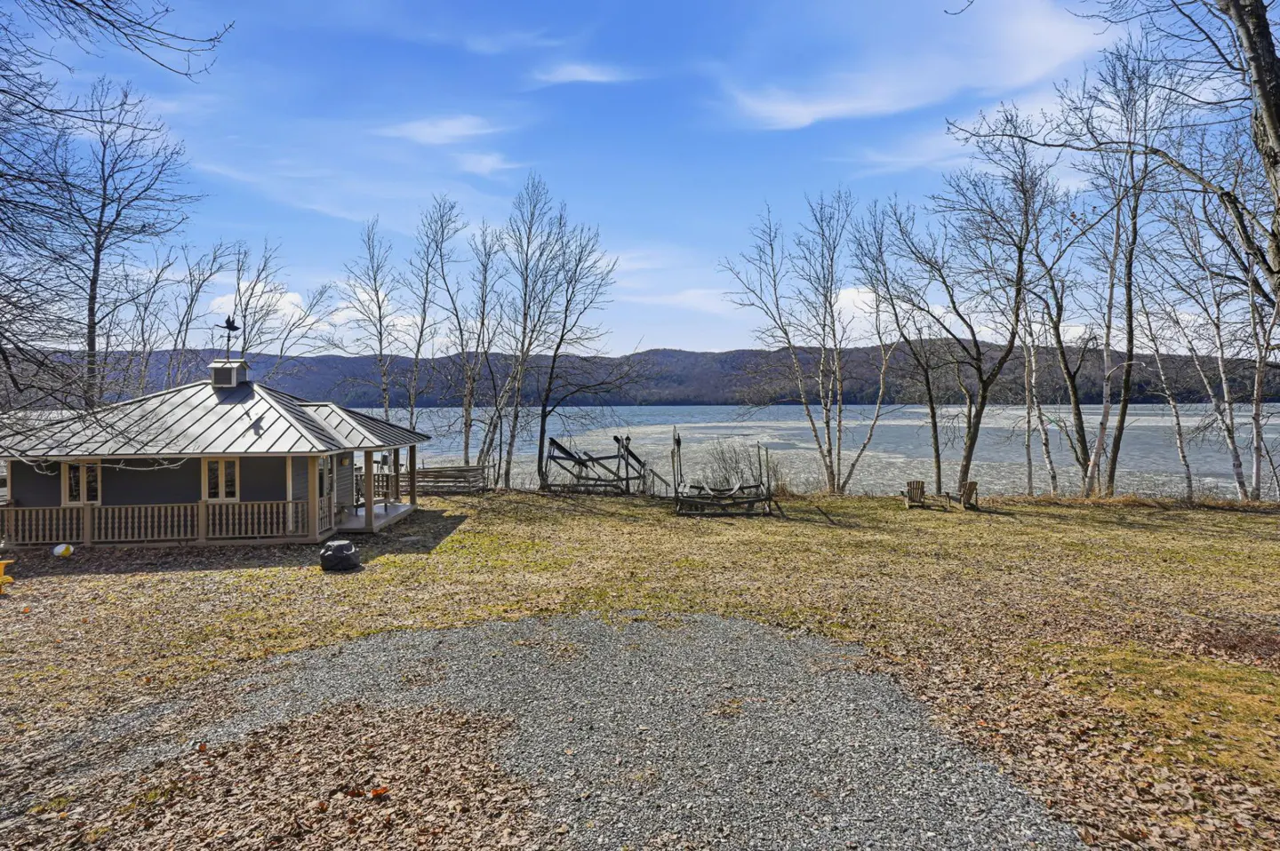 Lakefront property with a gray gazebo, bare trees, and a view of the water and distant mountains under a blue sky.