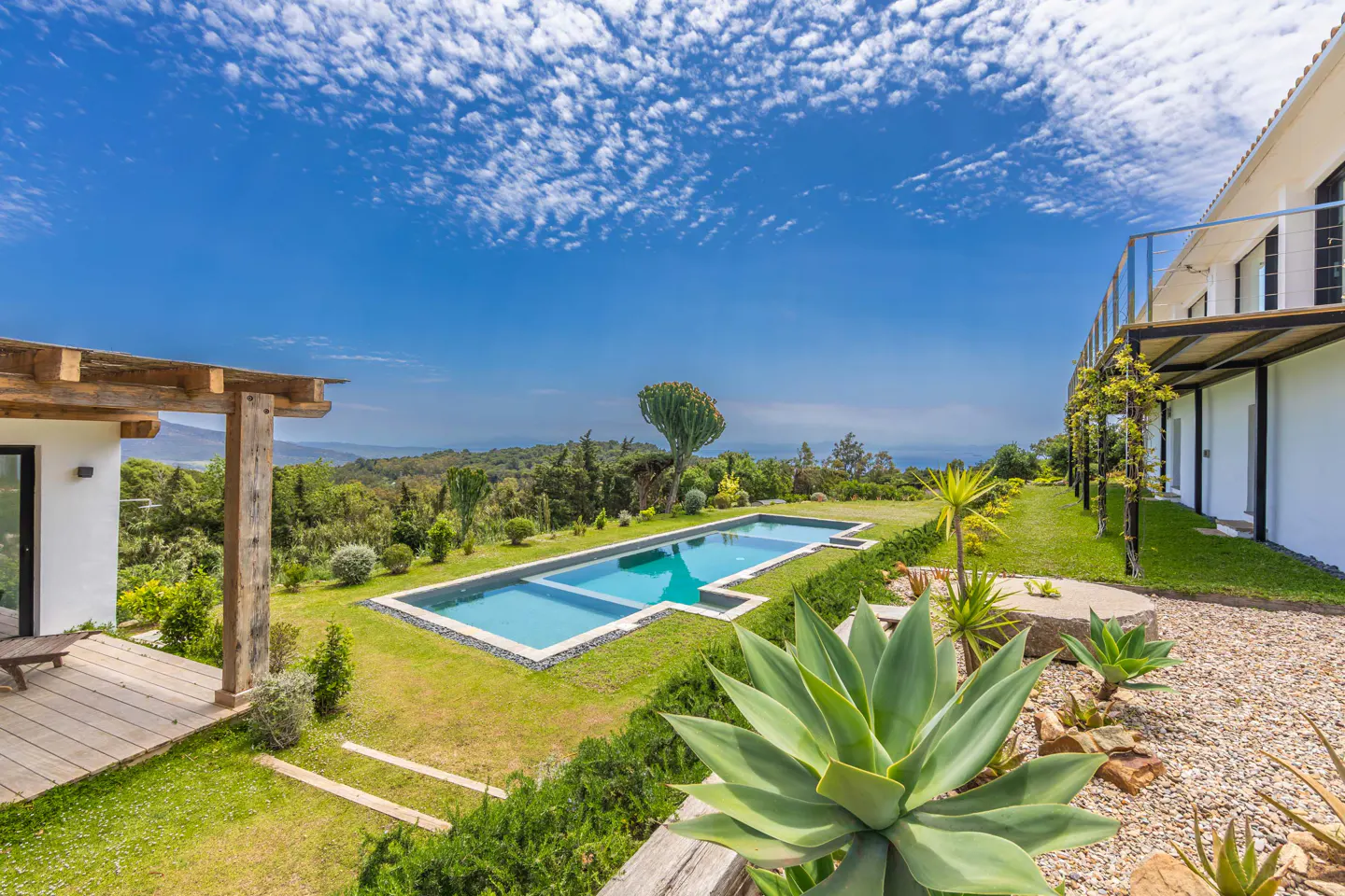 A luxurious backyard with a pool, green lawn, and lush vegetation under a bright blue sky with scattered clouds.