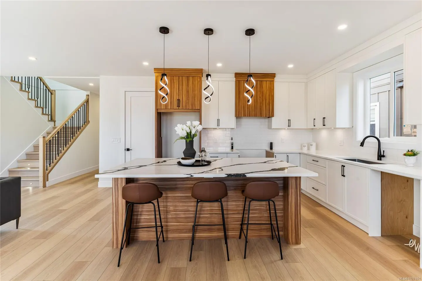 Bright kitchen with white cabinets, wood accents, and marble island. Three brown stools sit at the island, lit by spiral pendant lights. Stairs visible.
