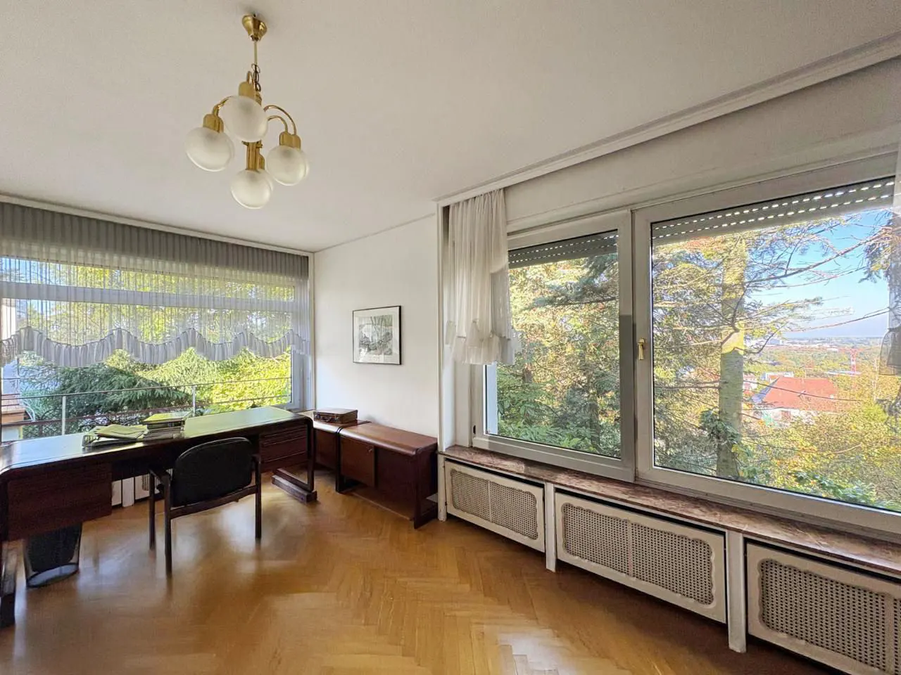 A bright office with a wood floor, a desk with a black chair, and a large window with a view of trees.
