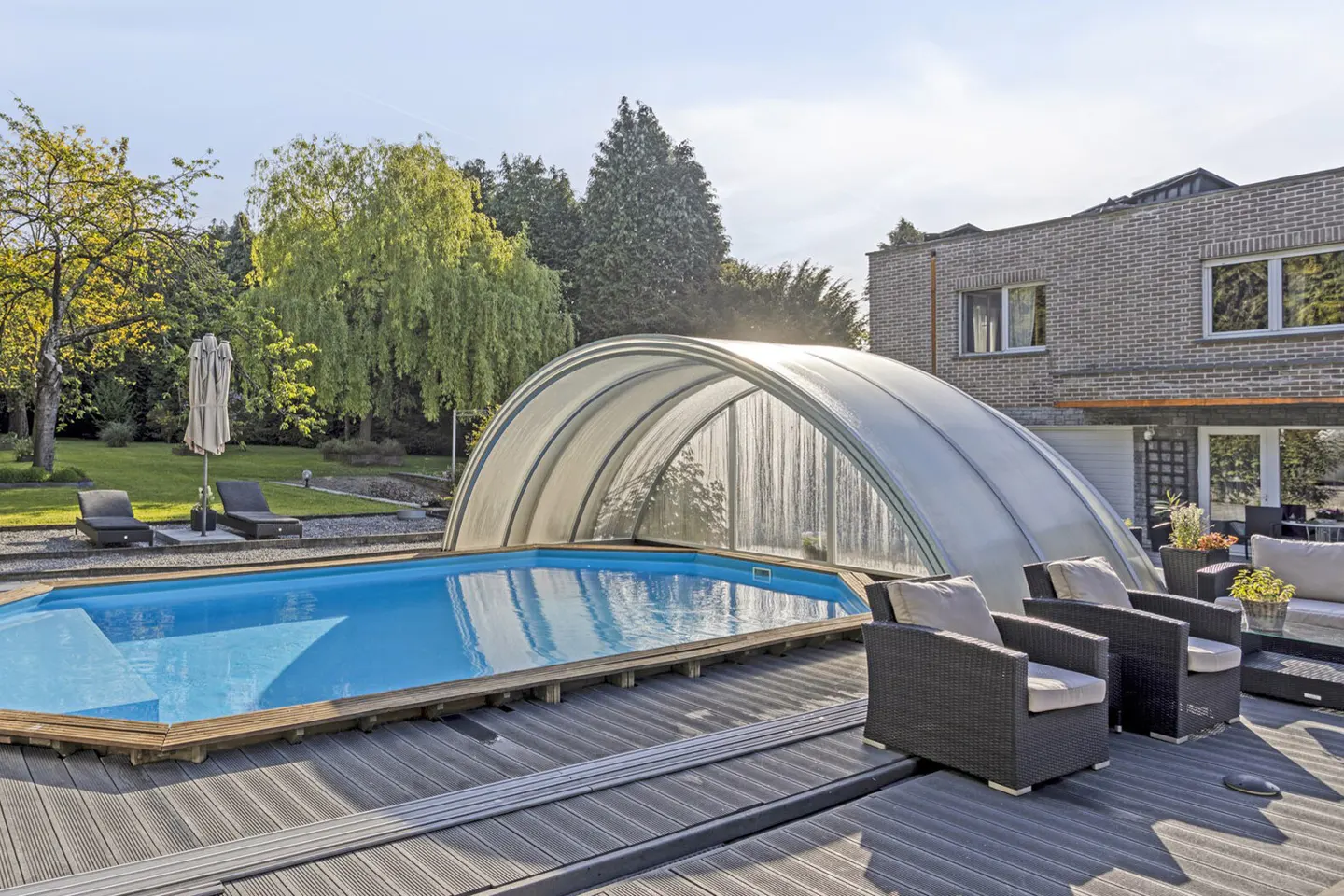 Outdoor pool with a retractable cover, surrounded by a wooden deck with lounge chairs and a gray brick house in the background.