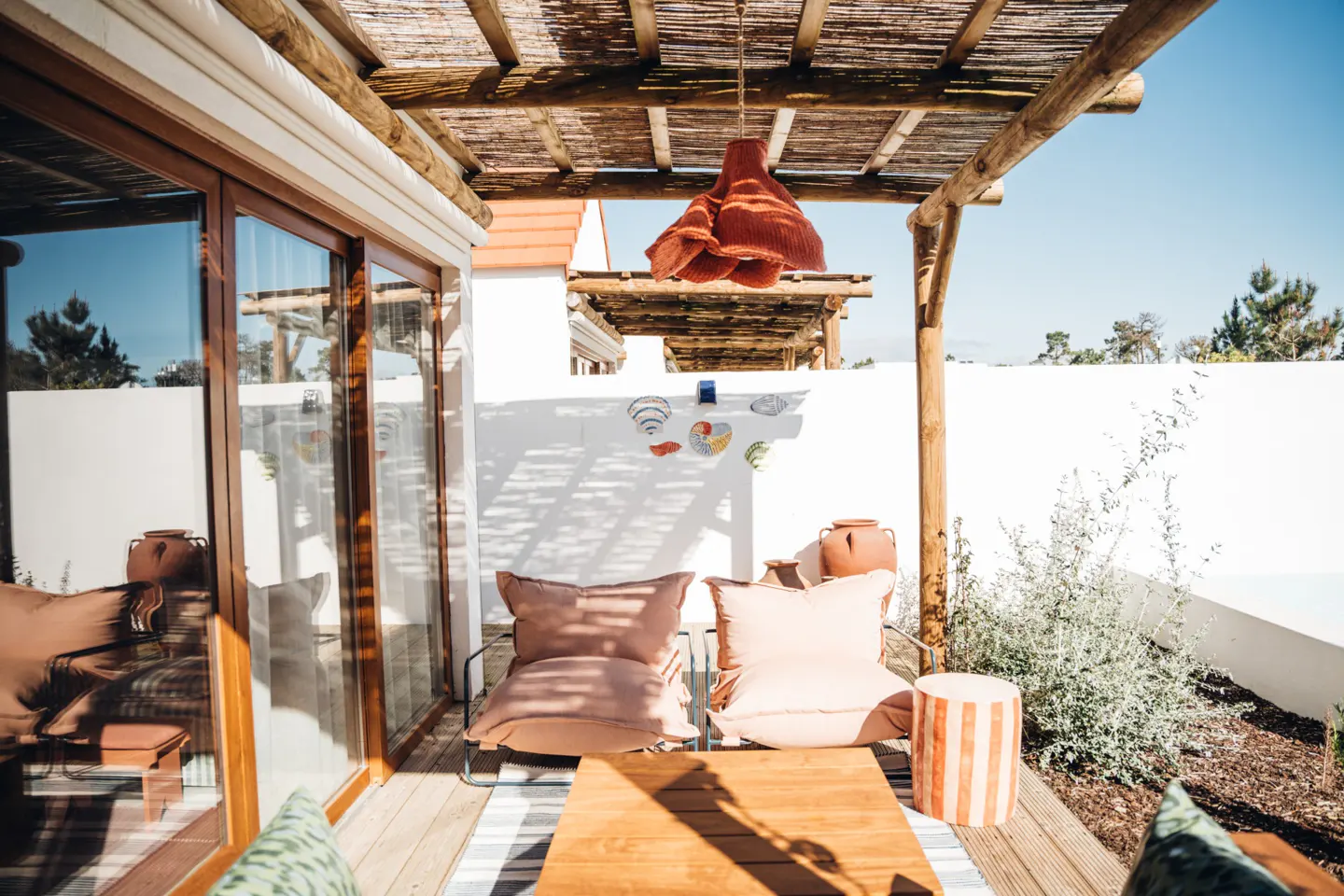 Outdoor patio with two chairs, a wooden table, and a woven rug. A terracotta lamp hangs from a thatched pergola. White walls and greenery surround the space.
