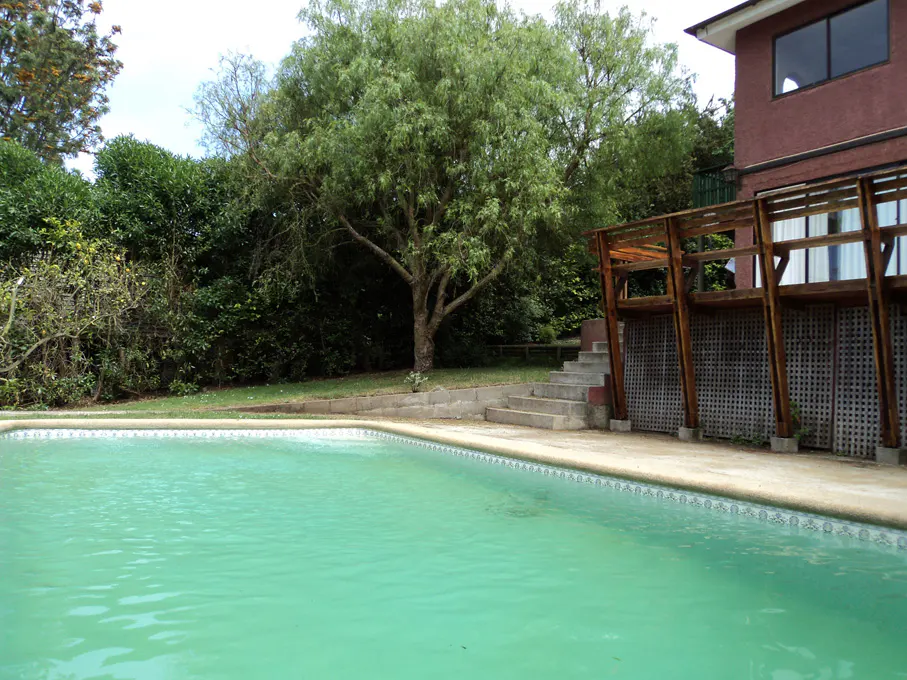 Outdoor pool with turquoise water, surrounded by green grass, trees, and a red house with a wooden deck.