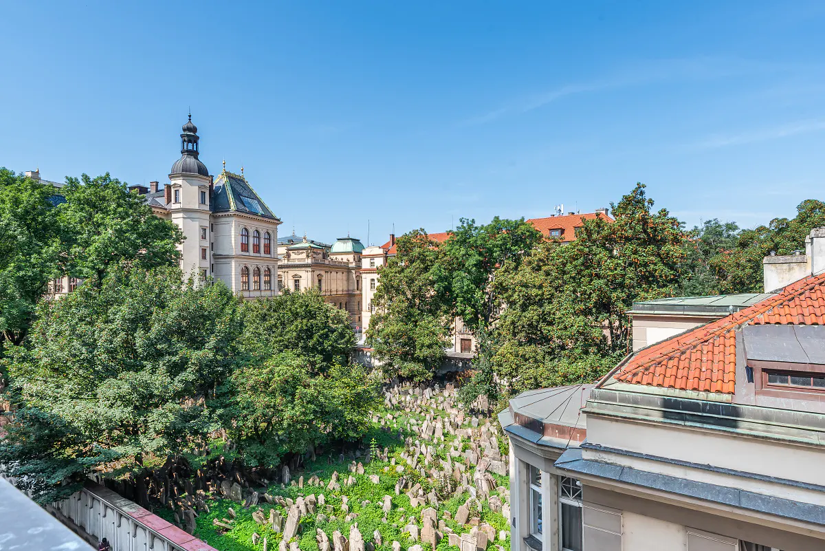 View of Prague's Old Jewish Cemetery, with headstones, trees, and buildings under a clear blue sky.