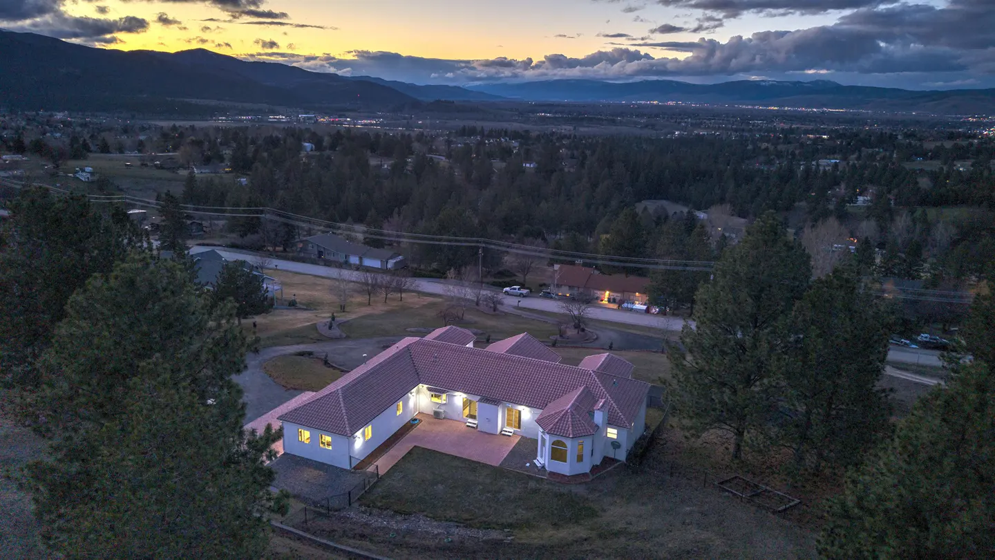 Aerial view of a white house with a red tile roof, surrounded by trees, with mountains in the background at dusk.