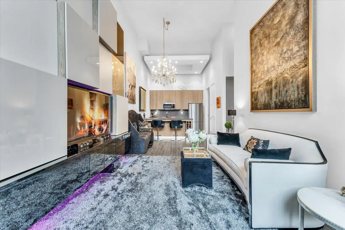 Living room with a fireplace, white sofa, and gray rug. A kitchen is visible in the background.