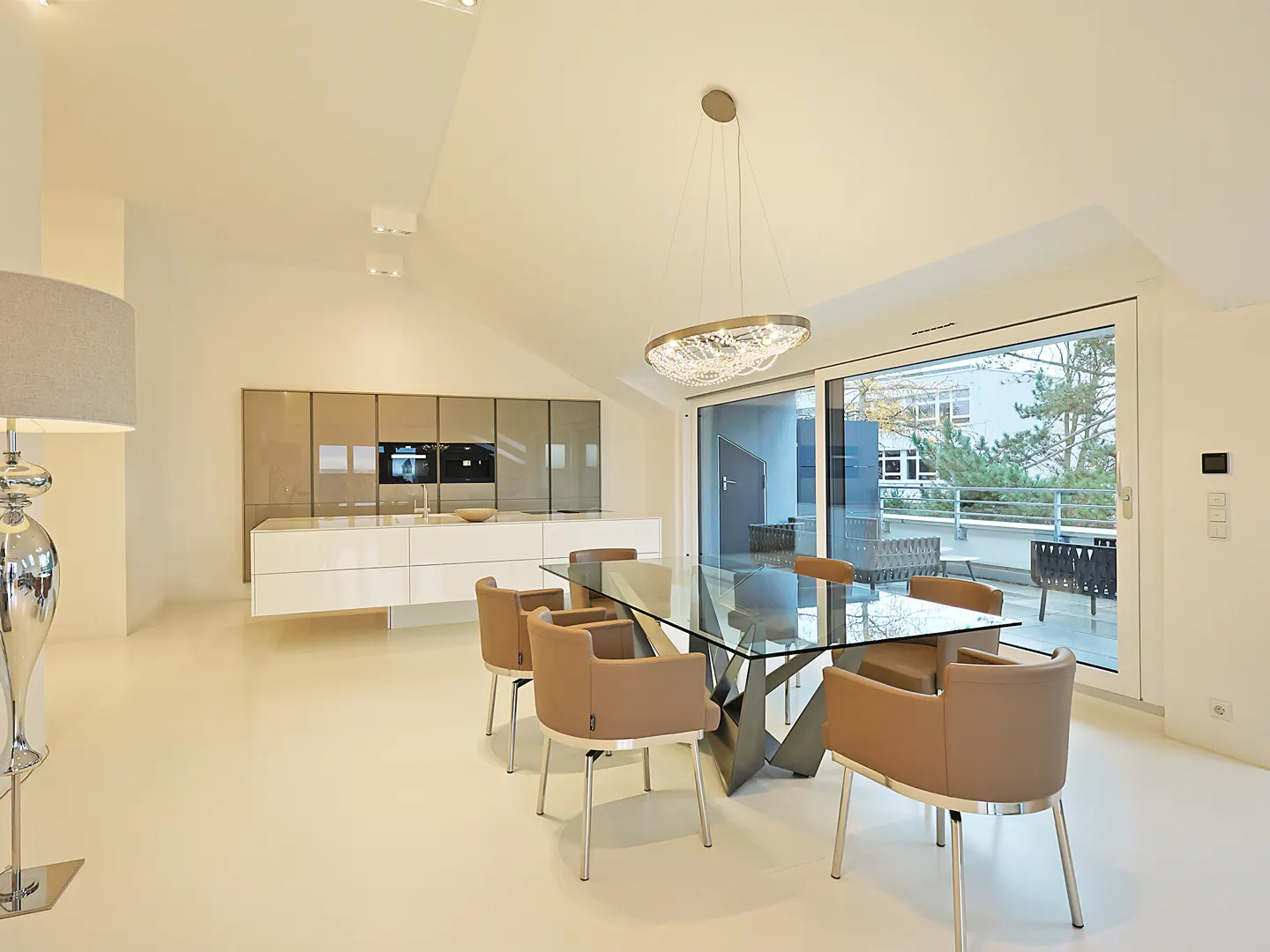Bright, modern dining room with glass table, tan chairs, and a crystal chandelier. Sliding glass doors lead to a balcony. Kitchen in background.