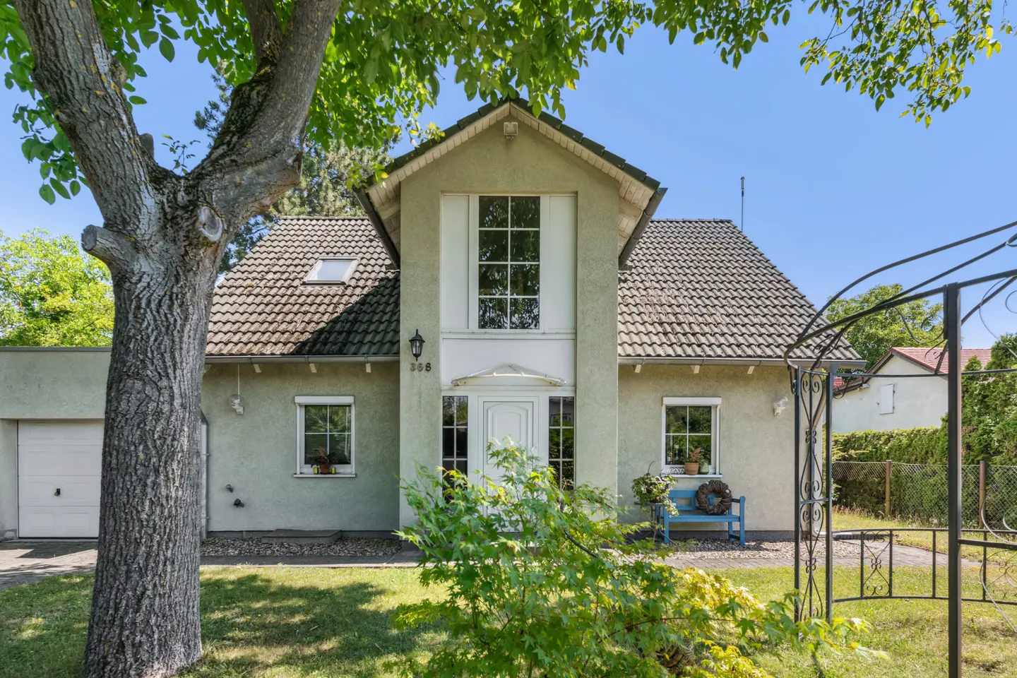 Two-story light green house with a gray tiled roof, white trim, and a large tree in the front yard.