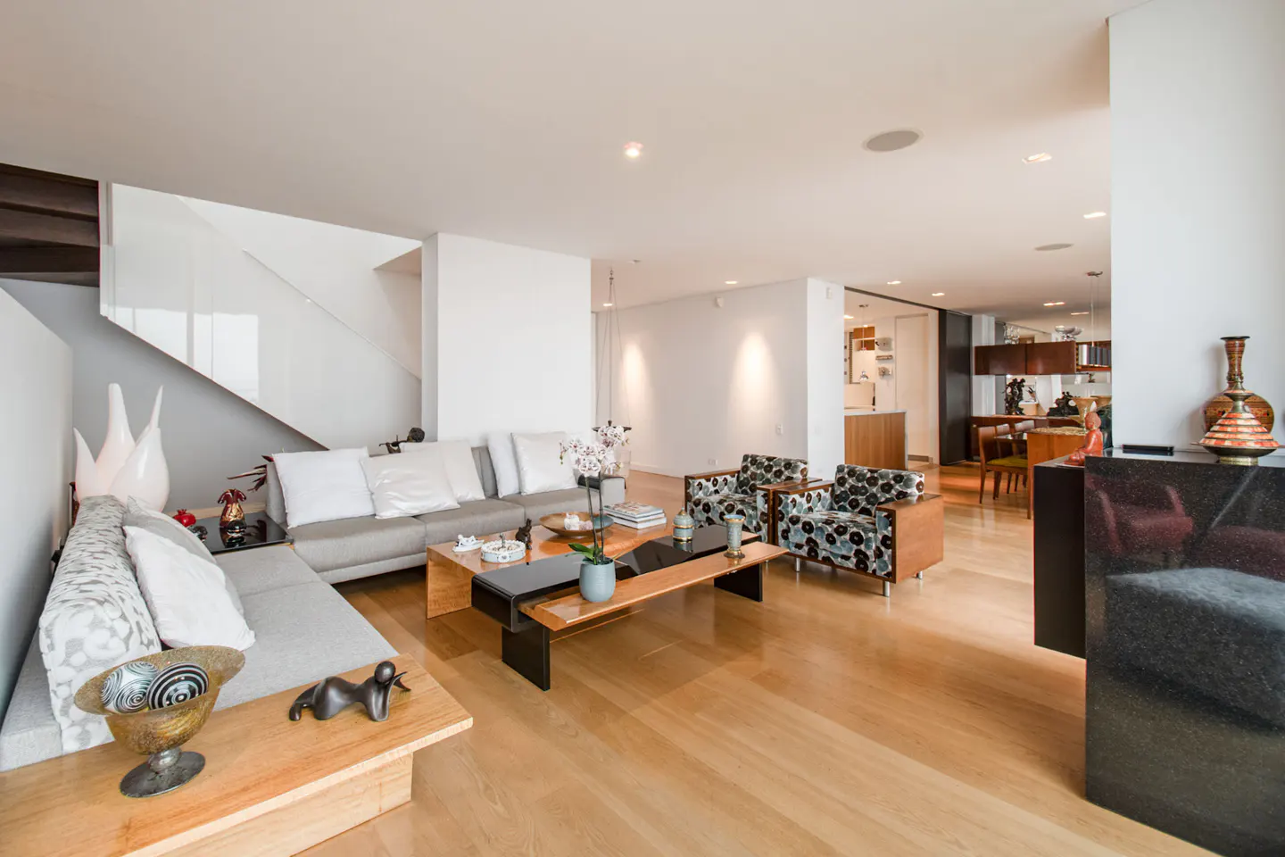 Bright, modern living room with light wood floors, gray sectional sofa, patterned armchairs, and a unique wooden coffee table. White walls and staircase.