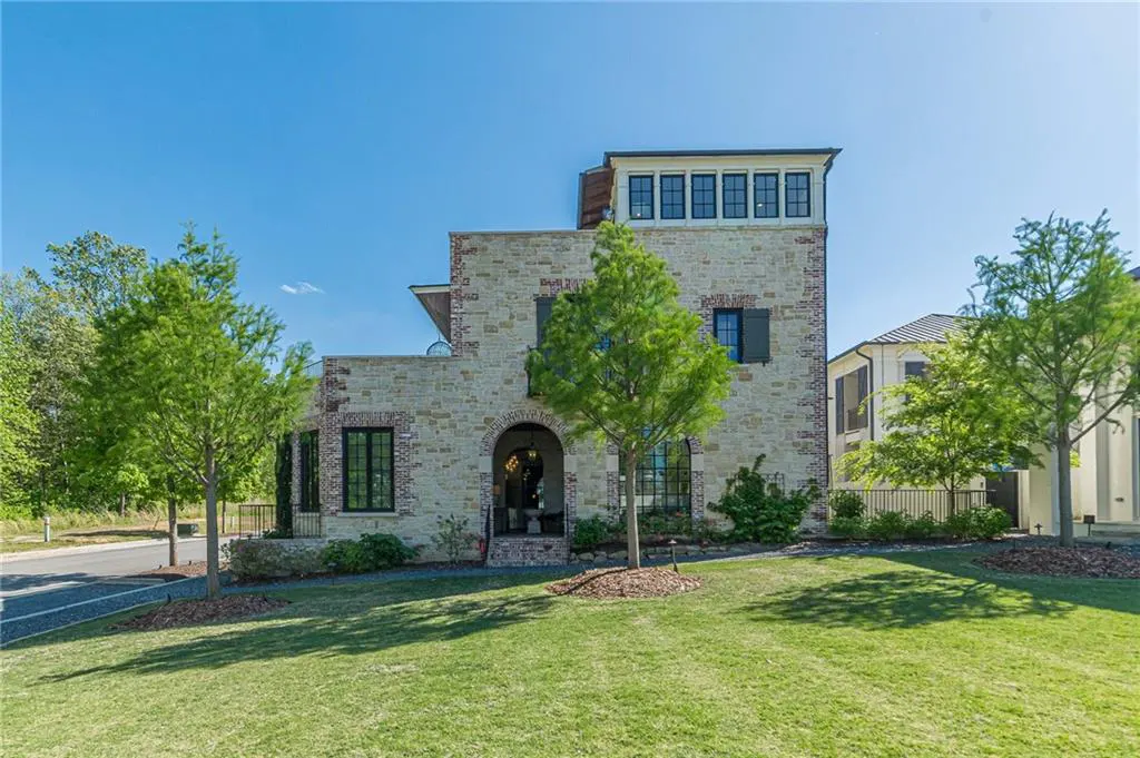 Three-story stone house with brick accents, arched entryway, and large windows, set on a green lawn under a clear blue sky.