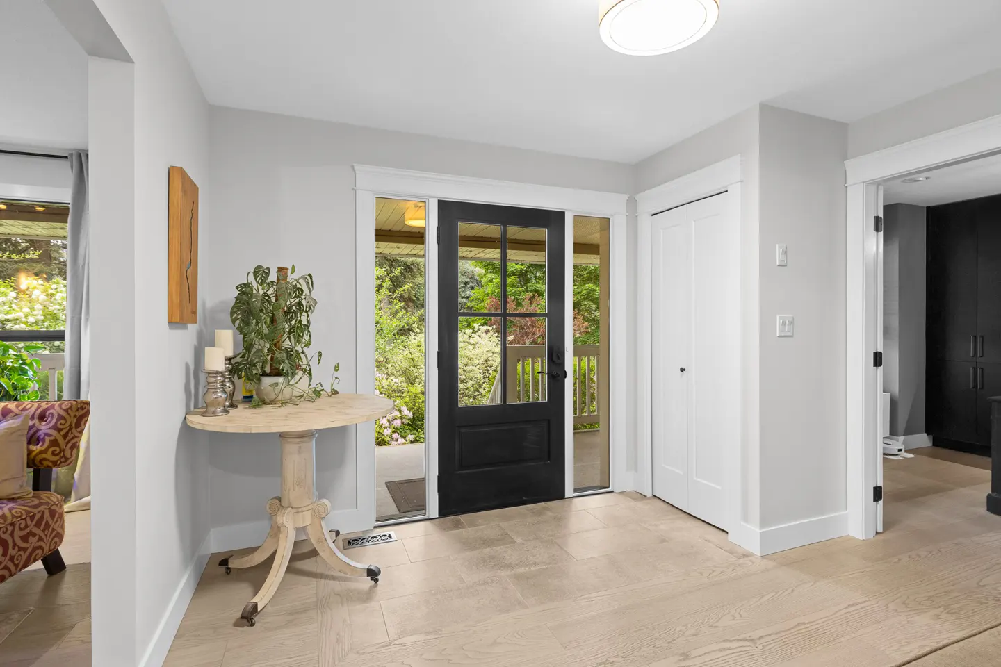 Bright entryway with light wood floors, a round pedestal table, a potted plant, and a black front door with glass panels.