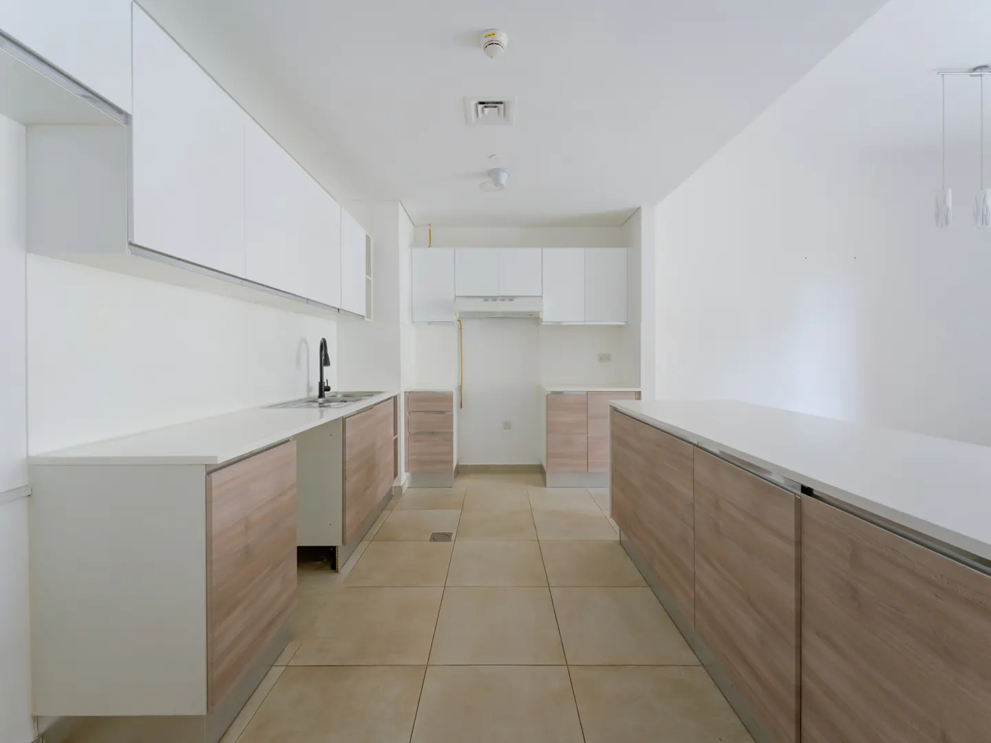 Bright, empty kitchen with white cabinets and light wood-grain lower cabinets. Beige tile floor and white walls.