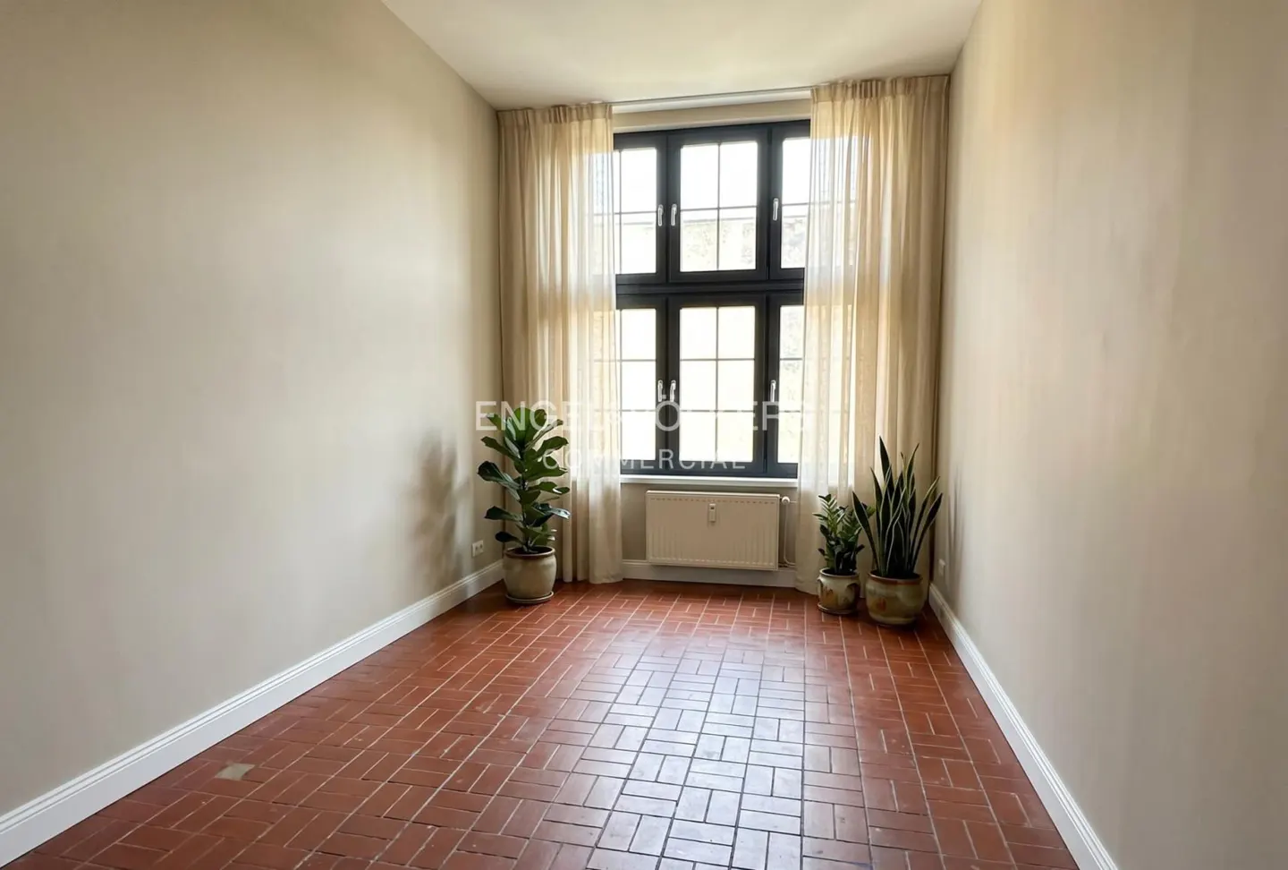 Empty room with brick floors, beige walls, and a black-framed window with sheer curtains. Potted plants sit near the window.