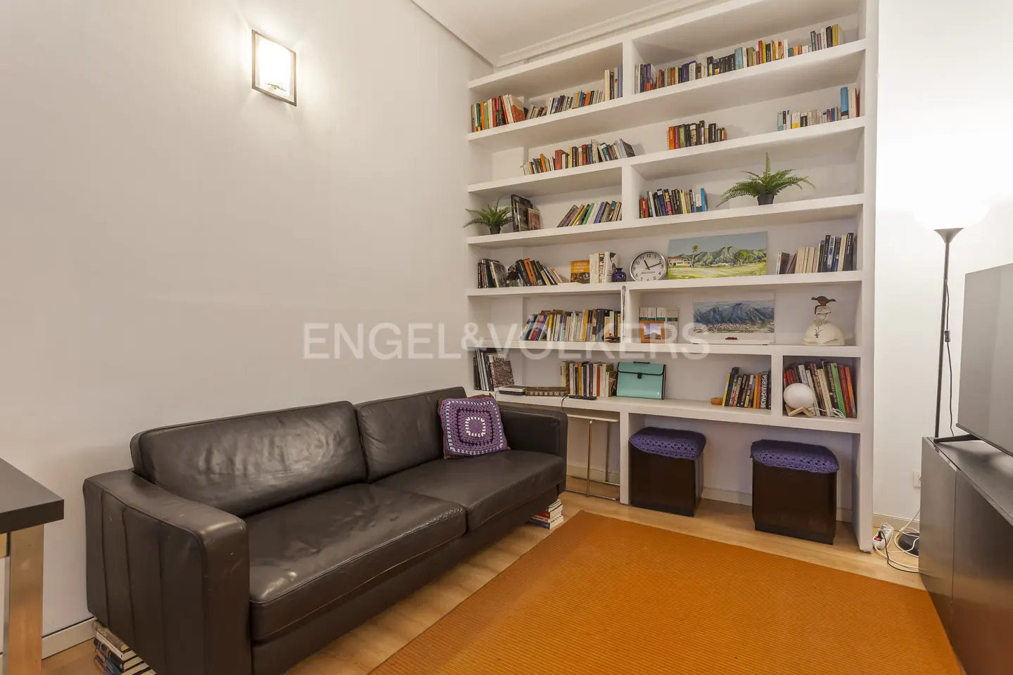 A dark leather sofa sits before a large white built-in bookshelf filled with books, decor, and two small purple stools.