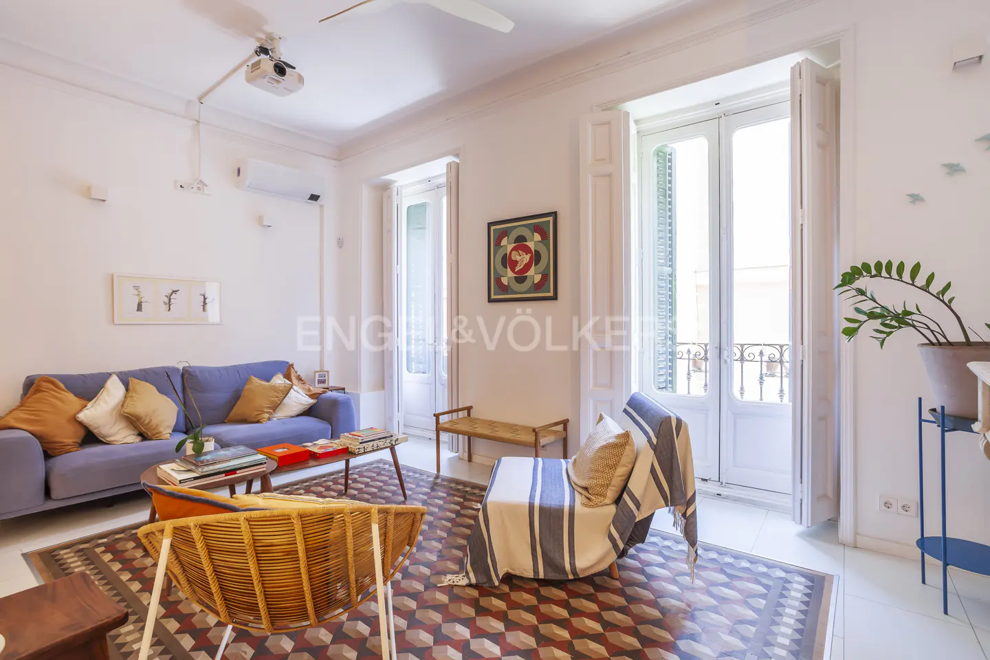 Bright living room with blue sofa, wicker chair, and patterned rug. White walls, French doors, and a projector on the ceiling.