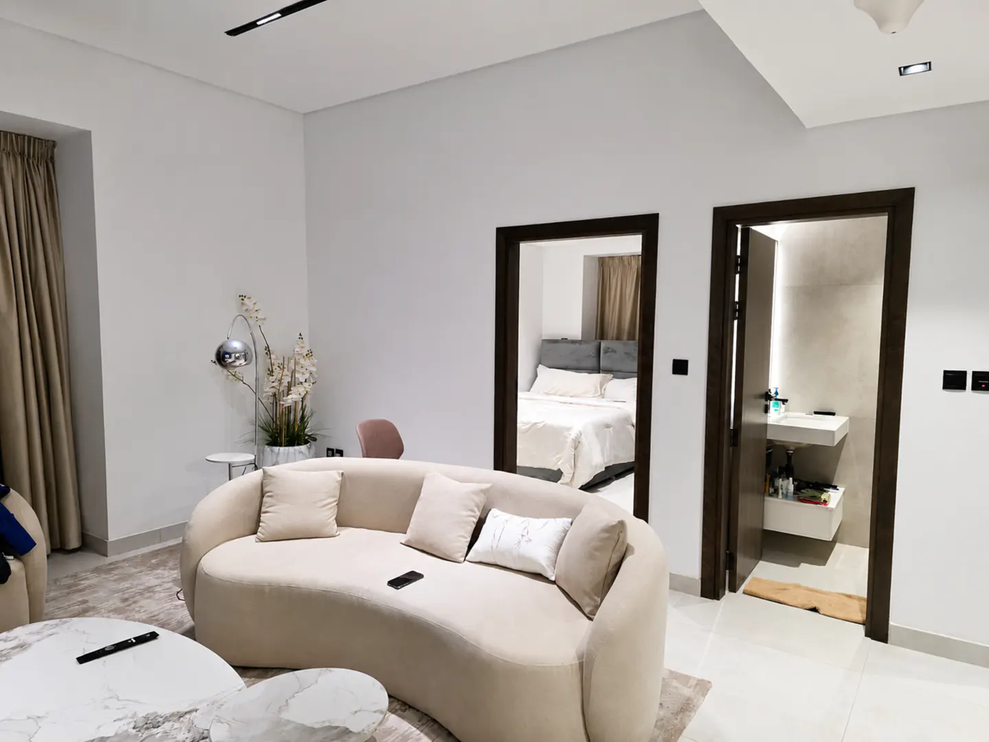 Living room with a beige curved sofa, marble coffee table, and views into a bedroom and bathroom through dark wood doorways.