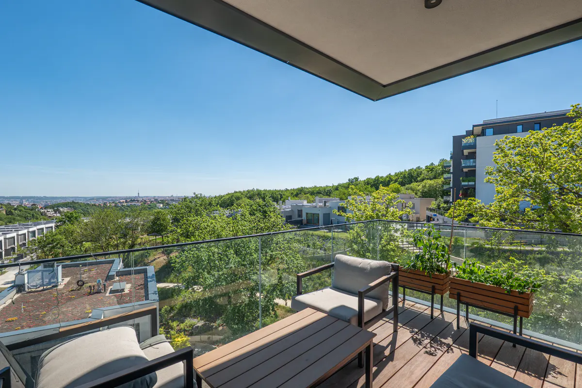 Modern balcony with wooden flooring, two armchairs, a coffee table, and planters overlooking a green landscape and city view.
